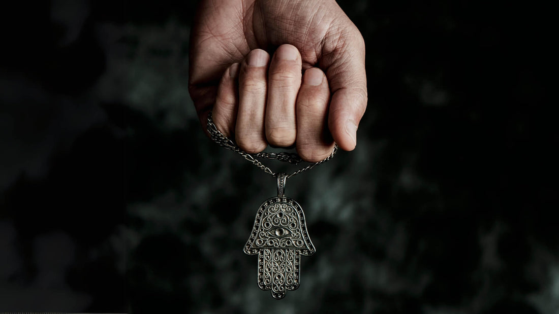 Hands held together around a symbolic silver Hamsa hand pendant on a dark background